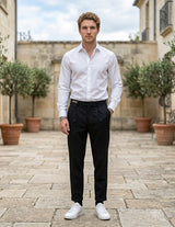 Male model wearing black pleated tailored pants with a white long-sleeve shirt in an outdoor stone walkway setting.