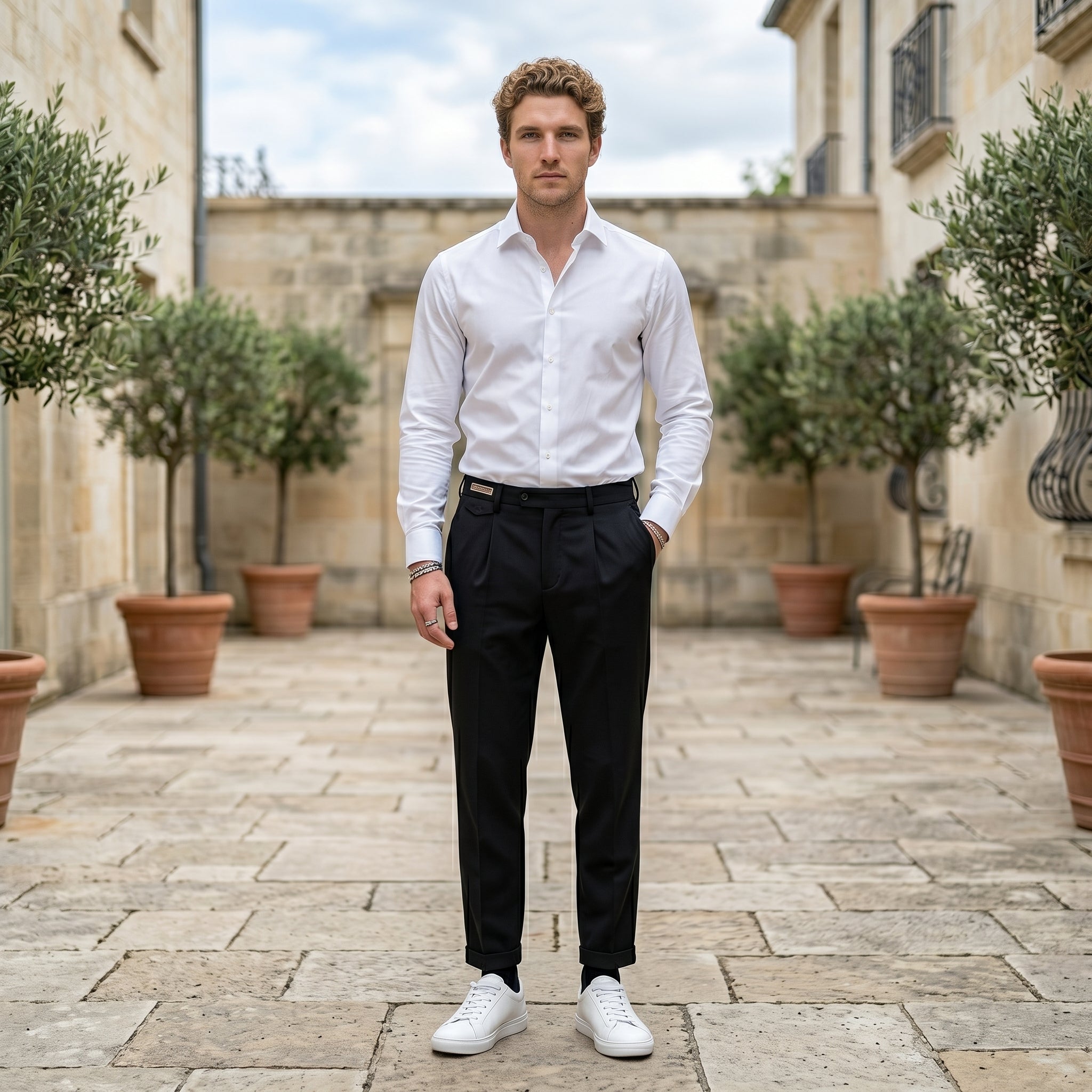 Male model wearing black pleated tailored pants with a white long-sleeve shirt in an outdoor stone walkway setting.
