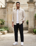 Male model wearing black relaxed-fit casual pants with a beige overshirt and white T-shirt in an outdoor stone walkway setting.