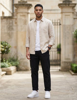 Male model wearing black relaxed-fit casual pants with a beige overshirt and white T-shirt in an outdoor stone walkway setting.