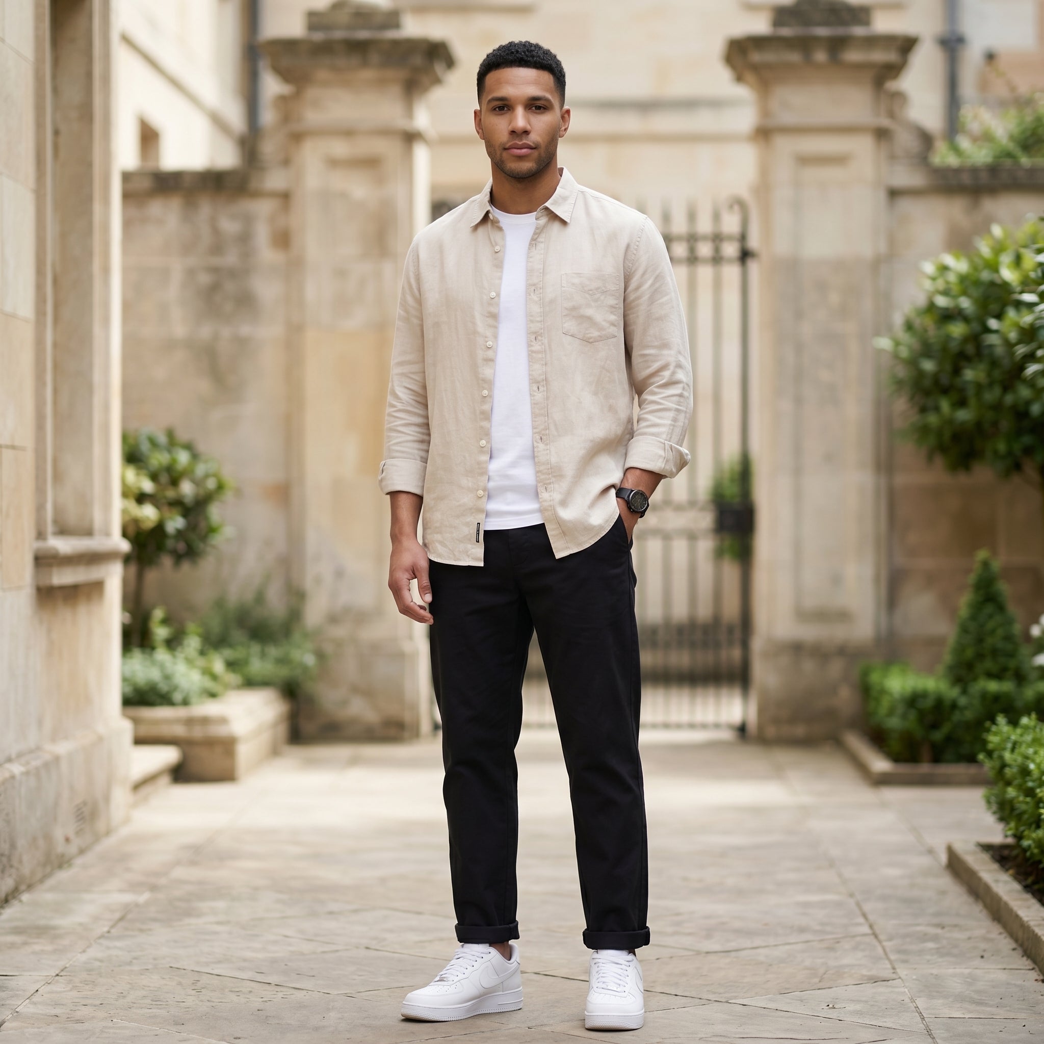 Male model wearing black relaxed-fit casual pants with a beige overshirt and white T-shirt in an outdoor stone walkway setting.