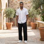Male model wearing black relaxed-fit casual pants with a white short-sleeve button-up shirt in an outdoor stone walkway setting.