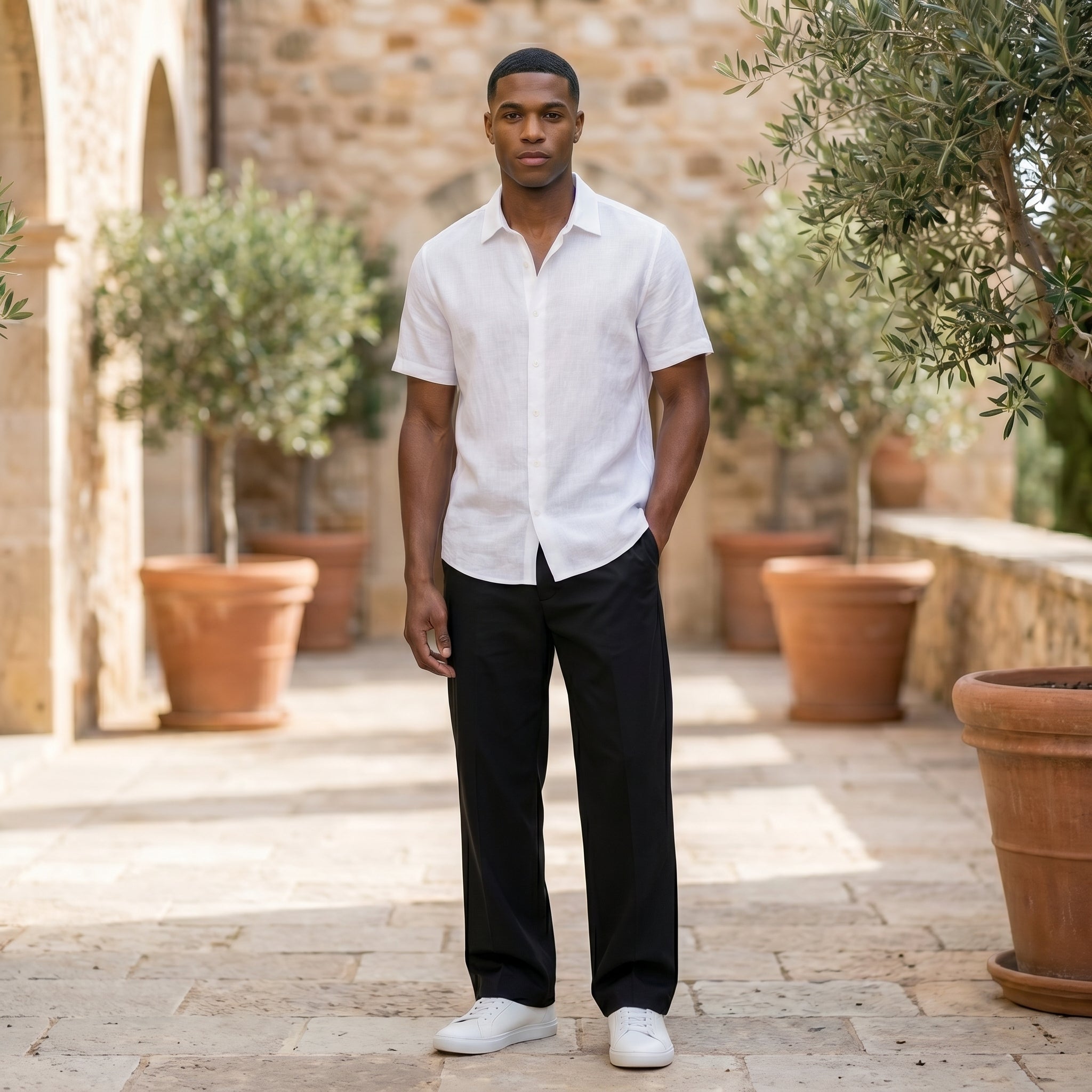 Male model wearing black relaxed-fit casual pants with a white short-sleeve button-up shirt in an outdoor stone walkway setting.