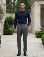 Male model wearing dark gray pleated tailored pants with a navy long-sleeve top in an outdoor walkway setting.
