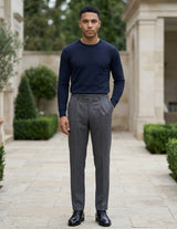Male model wearing dark gray pleated tailored pants with a navy long-sleeve top in an outdoor walkway setting.