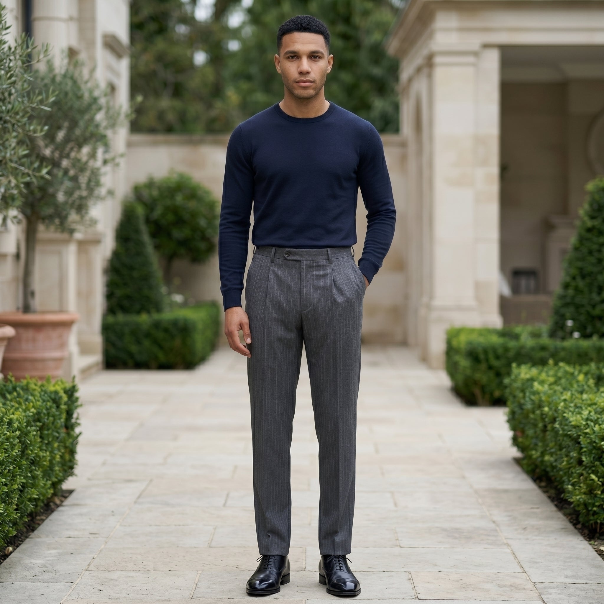 Male model wearing dark gray pleated tailored pants with a navy long-sleeve top in an outdoor walkway setting.
