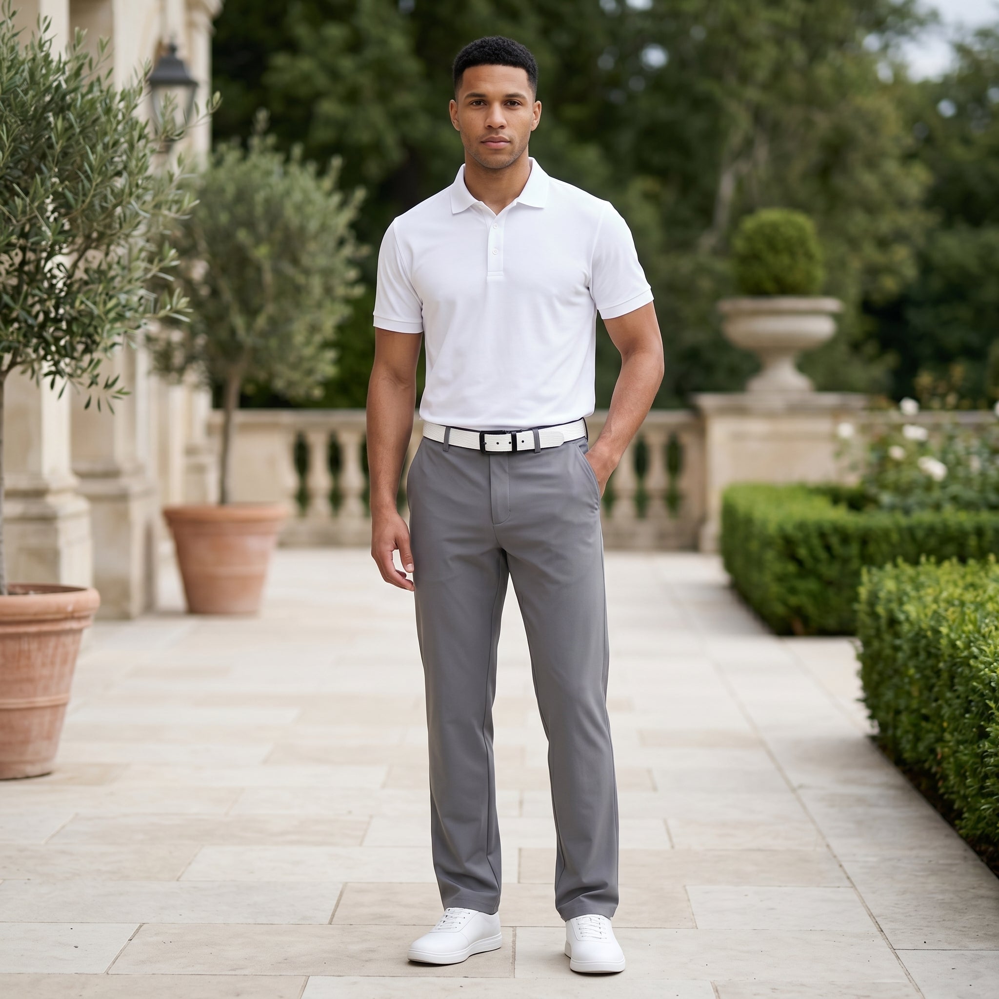 Male model wearing gray tailored casual pants with a white short-sleeve polo shirt in an outdoor garden walkway setting.