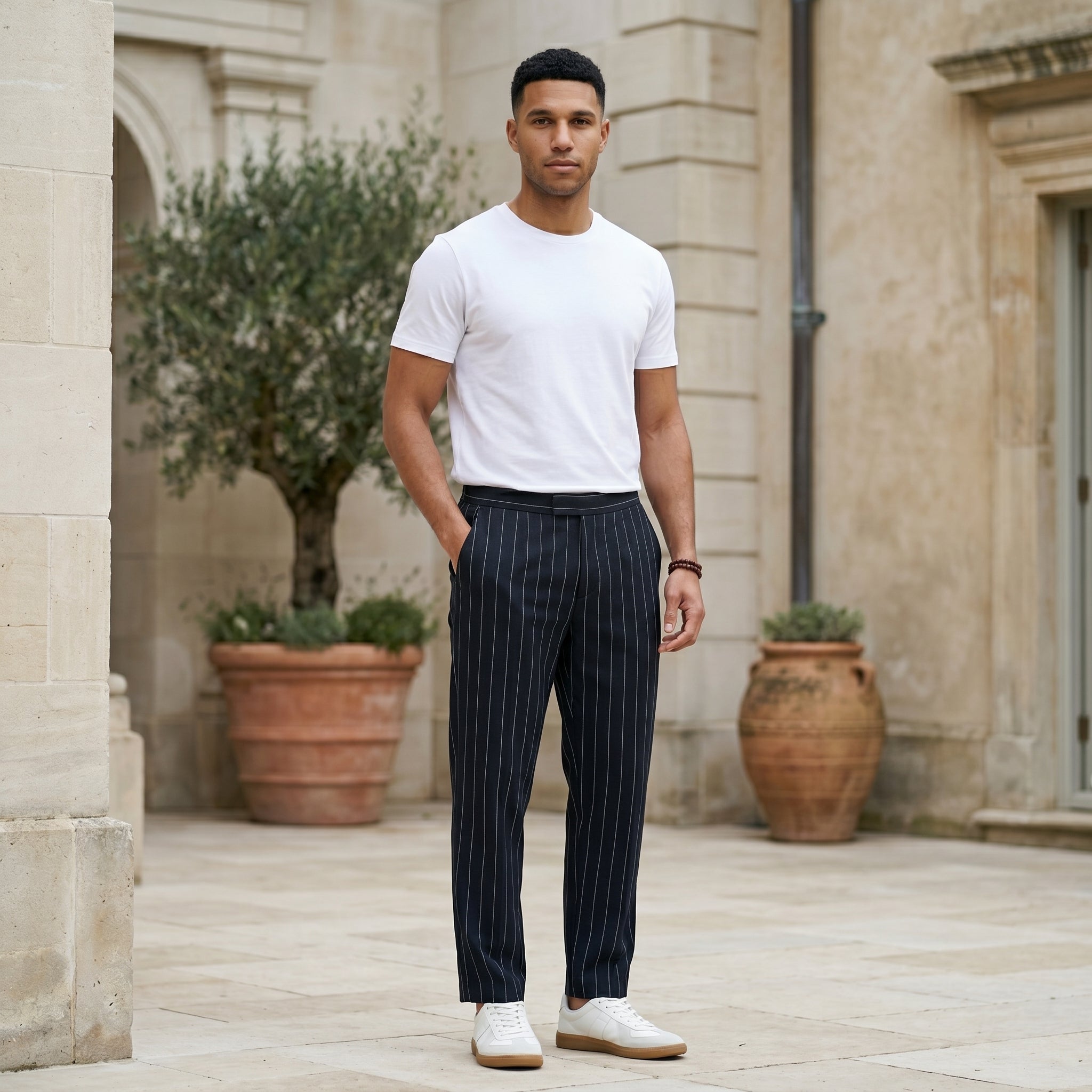 Male model wearing navy pinstripe relaxed-fit pants with a white T-shirt in an outdoor stone walkway setting.