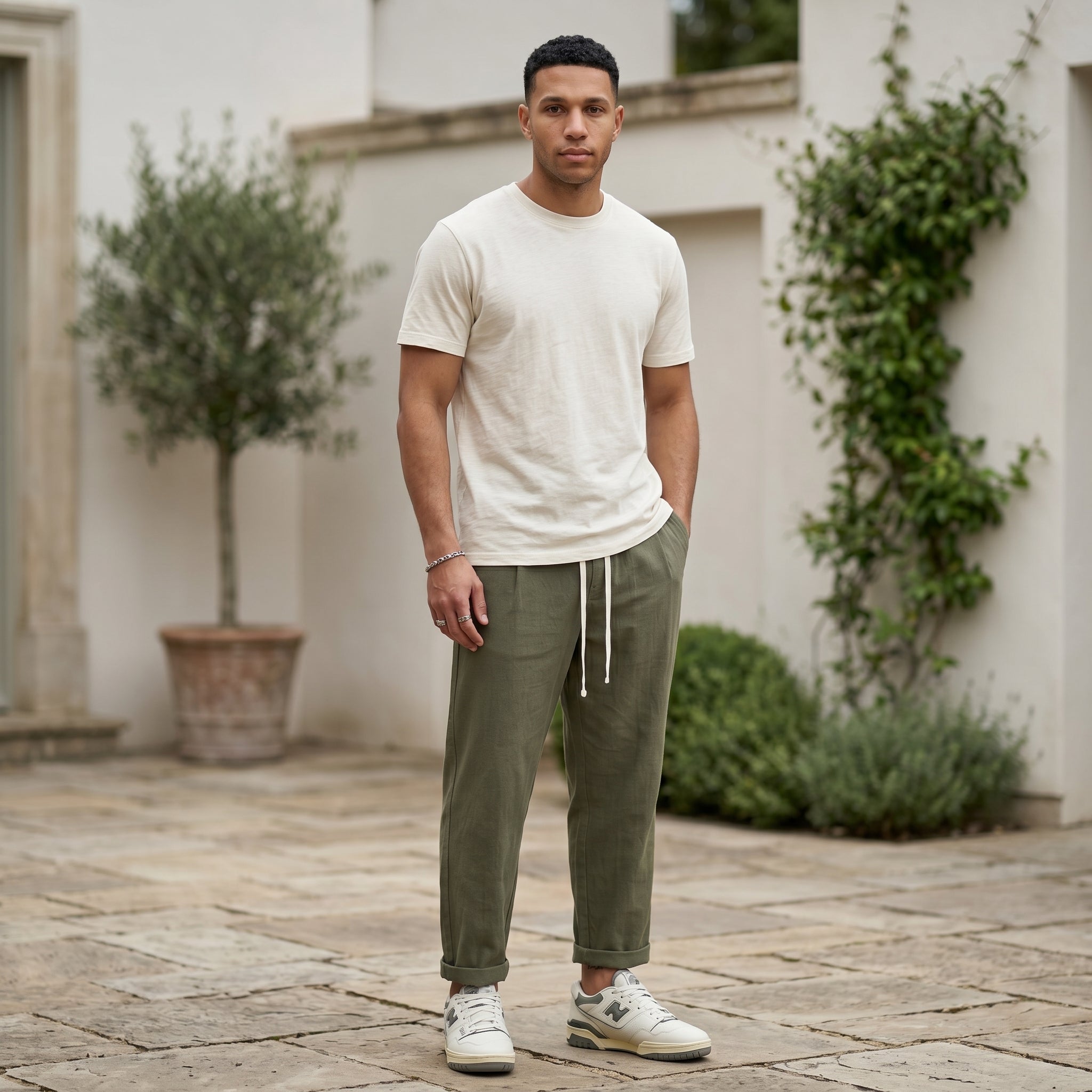 Male model wearing olive green drawstring jogger pants with a beige T-shirt in an outdoor stone walkway setting.