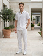 Male model wearing white drawstring relaxed-fit pants with a white short-sleeve button-up shirt in an outdoor walkway setting.