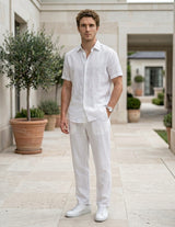 Male model wearing white drawstring relaxed-fit pants with a white short-sleeve button-up shirt in an outdoor walkway setting.