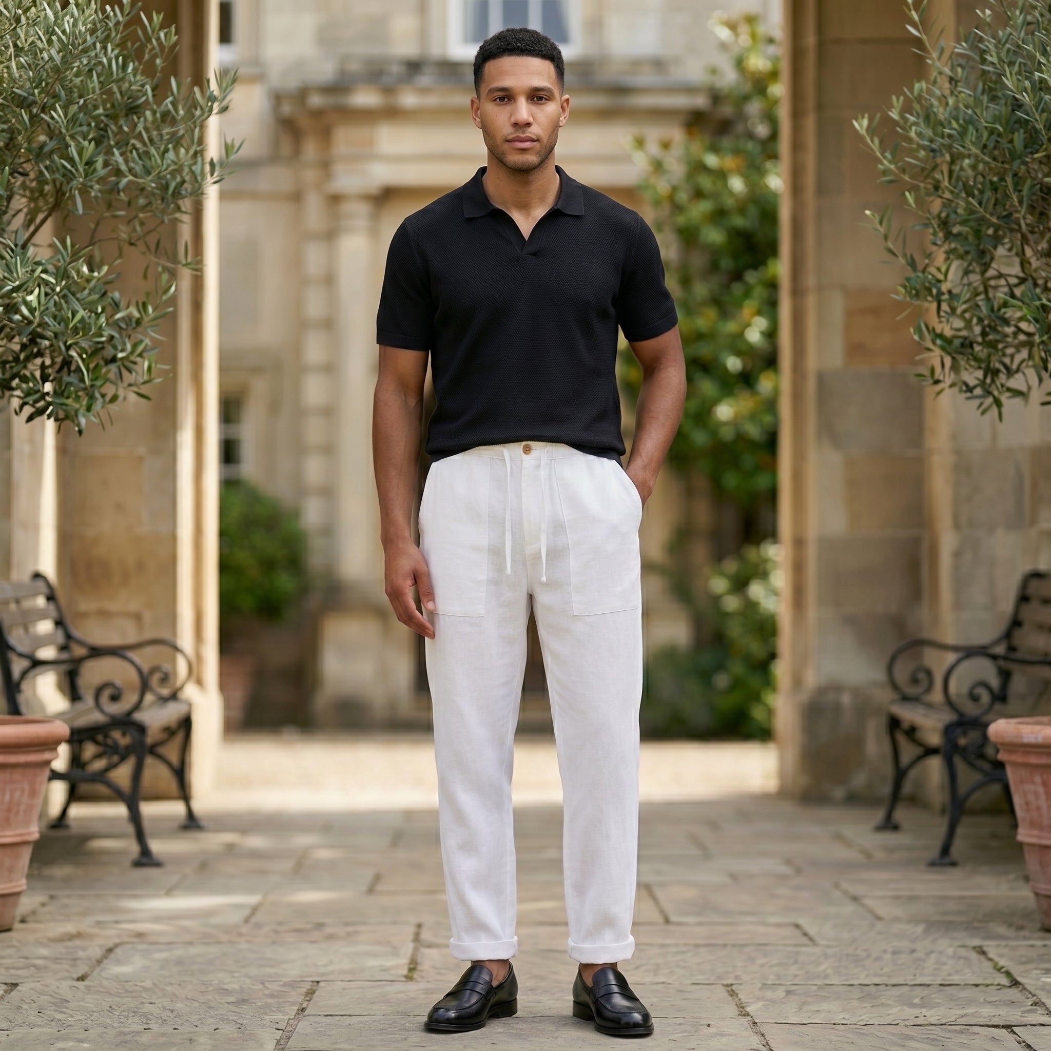 Male model wearing white relaxed-fit pants with a black short-sleeve polo shirt in an outdoor stone walkway setting.