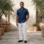 Male model wearing white relaxed-fit pants with a navy short-sleeve button-up shirt in an outdoor stone walkway setting.