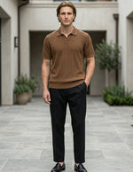 Man in a brown polo shirt and black pants standing in an outdoor setting with plants and buildings.