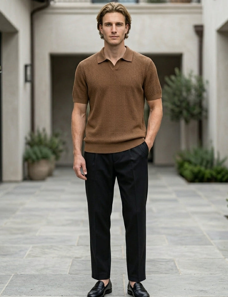 Man in a brown polo shirt and black pants standing in an outdoor setting with plants and buildings.