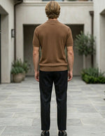 Man in a brown sweater and black pants standing in a courtyard with plants and buildings.