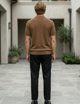 Man in a brown sweater and black pants standing in a courtyard with plants and buildings.