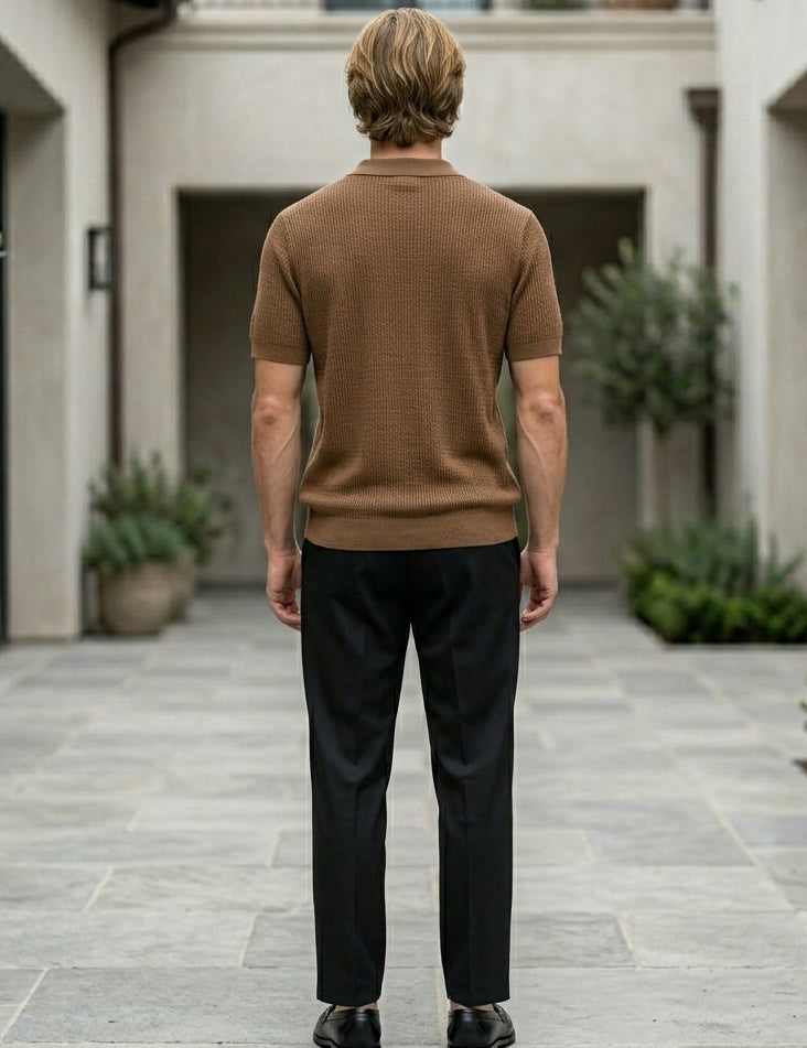Man in a brown sweater and black pants standing in a courtyard with plants and buildings.