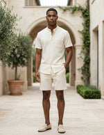 Man in a cream outfit standing in an outdoor setting with architectural elements and plants.