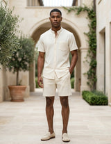 Man in a cream outfit standing in an outdoor setting with architectural elements and plants.