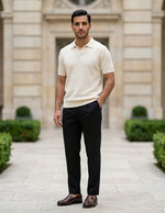 Man in a cream shirt and black pants standing in front of a building with greenery.