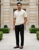 Man in a cream shirt and black pants standing in front of a building with greenery.