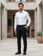 Man in a white shirt and black pants standing in an outdoor setting with plants.