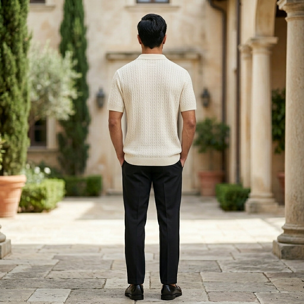 Man in a white sweater and black pants standing in a courtyard with columns and plants.