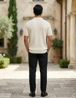Man in a white sweater and black pants standing in a courtyard with columns and plants.