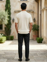 Man in a white sweater and black pants standing in a courtyard with columns and plants.