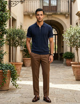 Man in navy polo shirt and brown pants standing in an outdoor setting with plants and architecture.