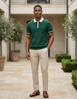 Man wearing a green polo shirt and beige pants standing in an outdoor setting with plants and a building.