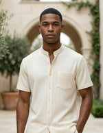Man wearing a beige button-up shirt standing in an outdoor setting with arches and plants.