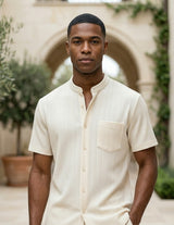 Man wearing a beige button-up shirt standing in an outdoor setting with arches and plants.