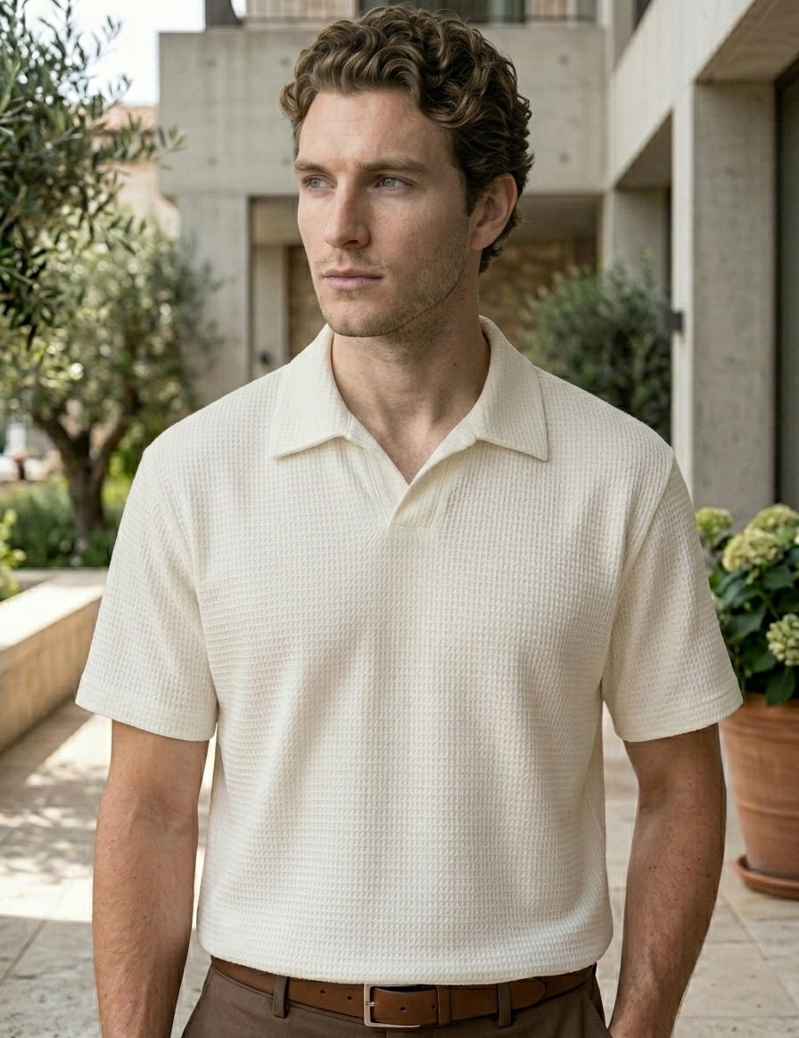 Man wearing a beige checkered polo shirt standing outdoors with greenery and a building in the background.