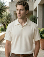 Man wearing a beige checkered polo shirt standing outdoors with greenery and a building in the background.