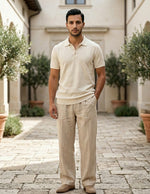 Man wearing a beige polo shirt and pants standing in an outdoor setting with stone pavement and greenery.