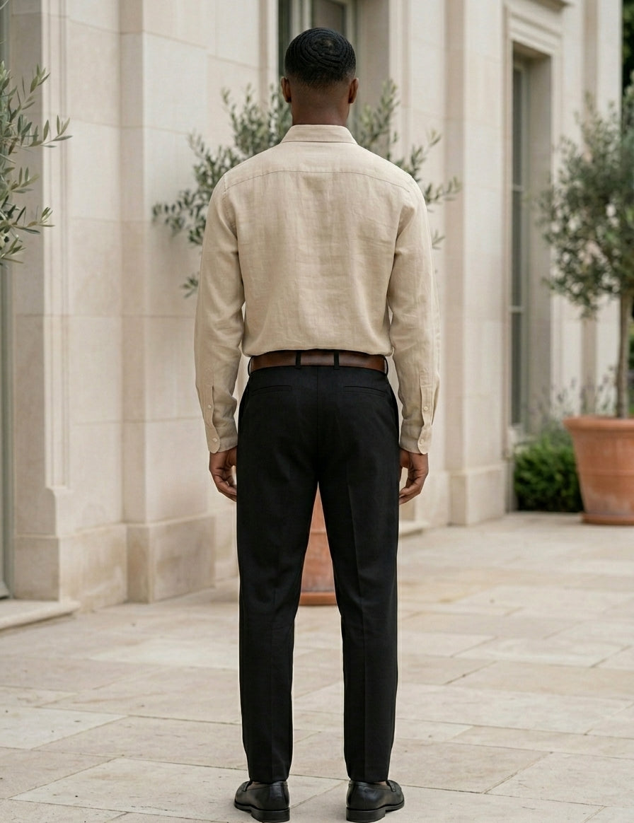 Man wearing a beige shirt and black pants standing in front of a stone building with plants.