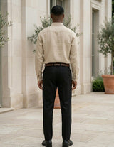 Man wearing a beige shirt and black pants standing in front of a stone building with plants.
