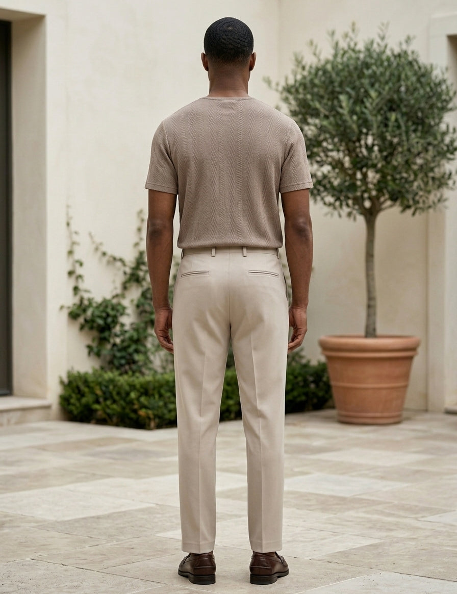 Man wearing a beige shirt and pants standing in an outdoor setting with plants and a building.