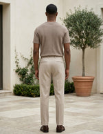 Man wearing a beige shirt and pants standing in an outdoor setting with plants and a building.