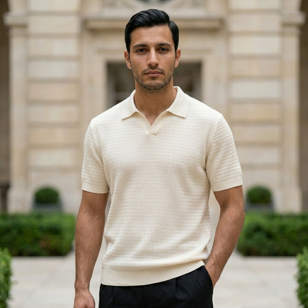Man wearing a beige short-sleeve knit shirt standing in front of a building with greenery.