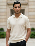Man wearing a beige short-sleeve knit shirt standing in front of a building with greenery.