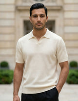 Man wearing a beige short-sleeve knit shirt standing in front of a building with greenery.