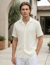 Man wearing a beige short-sleeve shirt and white pants standing outdoors.