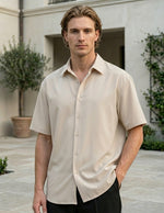 Man wearing a beige short-sleeve shirt standing outdoors with greenery in the background