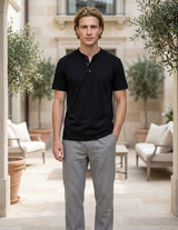 Man wearing a black polo shirt and gray pants standing in an outdoor setting with plants and furniture.