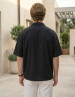 Man wearing a black polo shirt and white pants standing outdoors with plants and a building in the background.