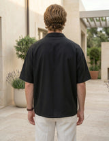 Man wearing a black polo shirt and white pants standing outdoors with plants and a building in the background.