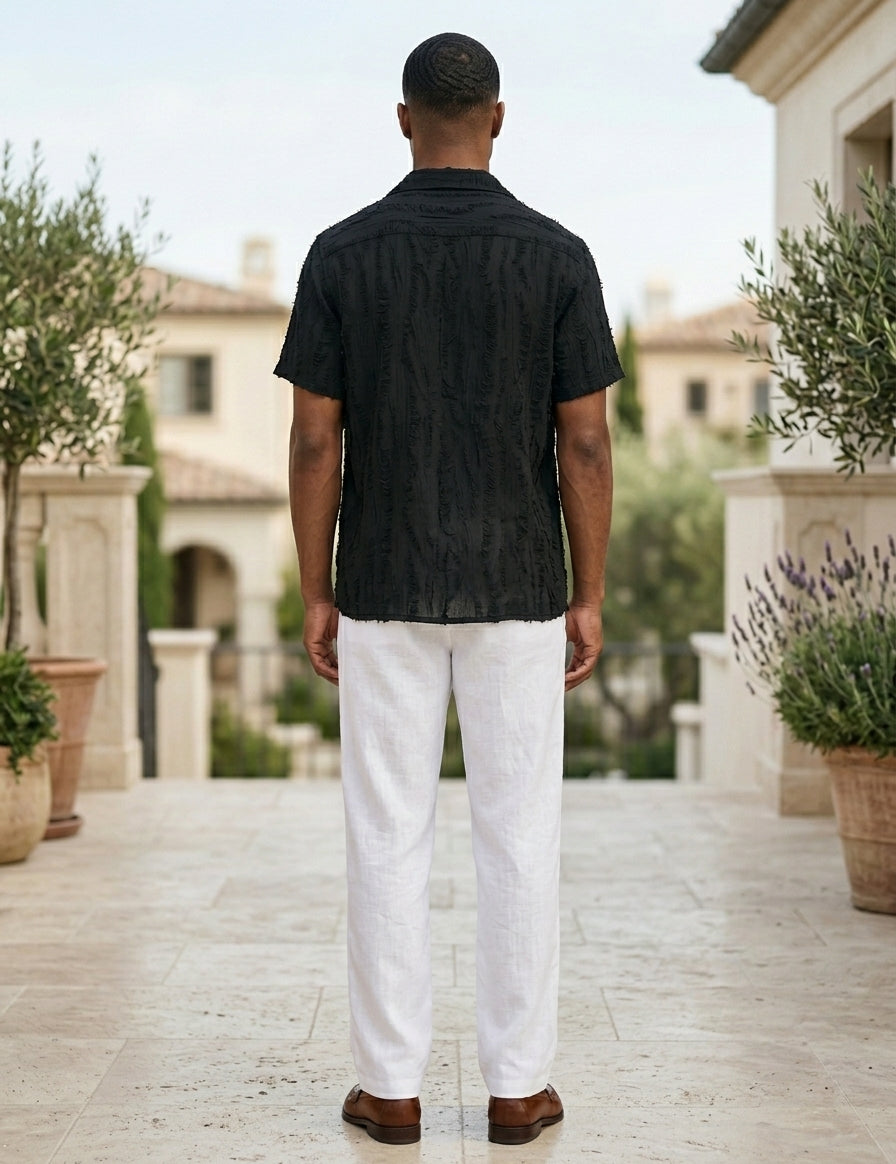 Man wearing a black shirt and white pants standing in an outdoor setting with plants and buildings in the background.